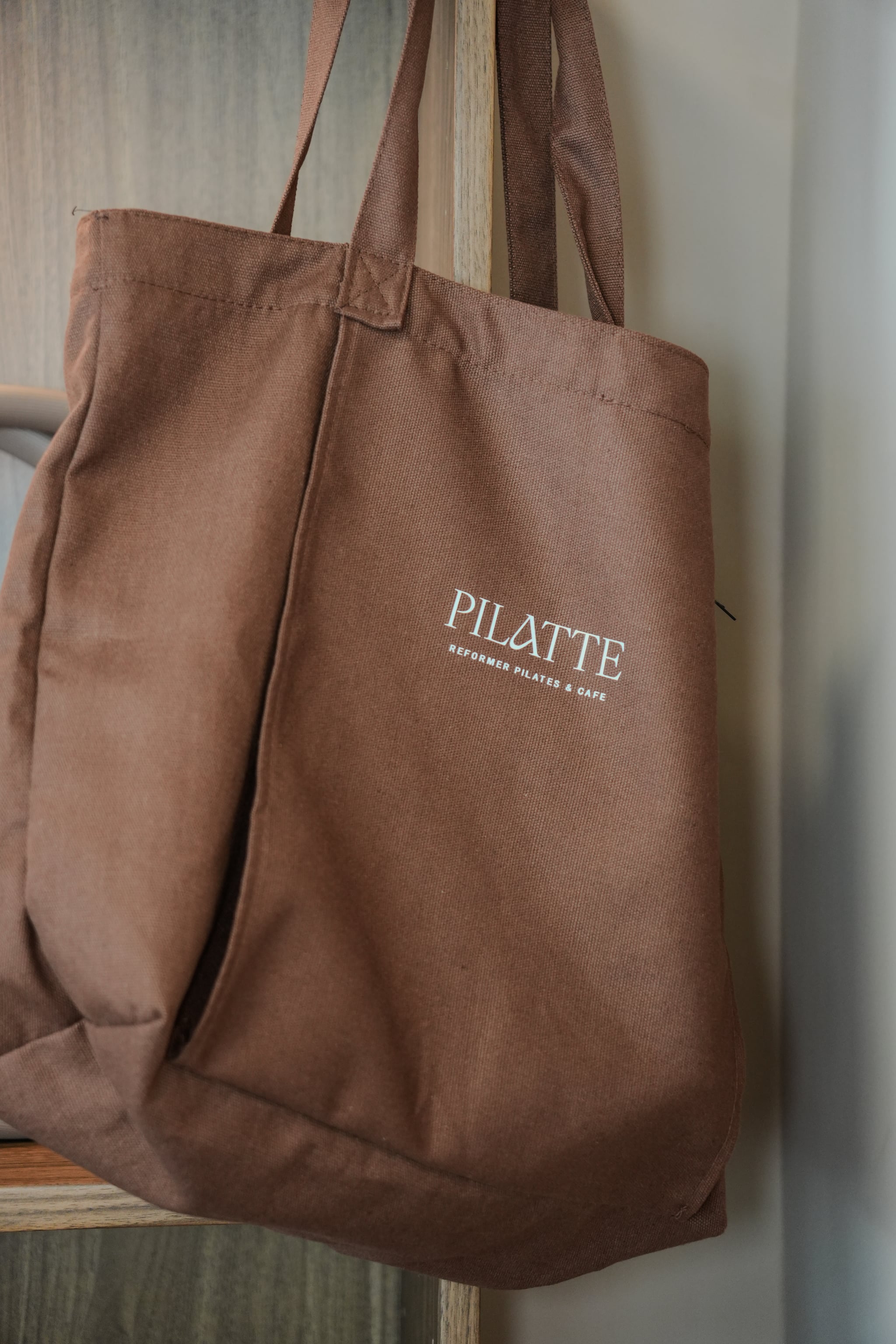 Braune Stofftasche mit Pilatte-Aufdruck, an einem Holzgestell hängend